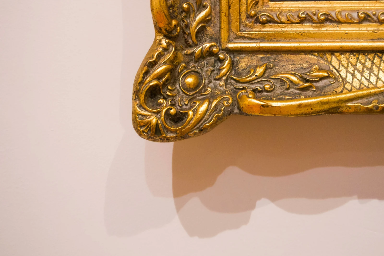 Close-up of the corner of an ornate gilded picture frame where the gold leaf has rubbed away to reveal the dull dark ground underneath, against a plain wall.