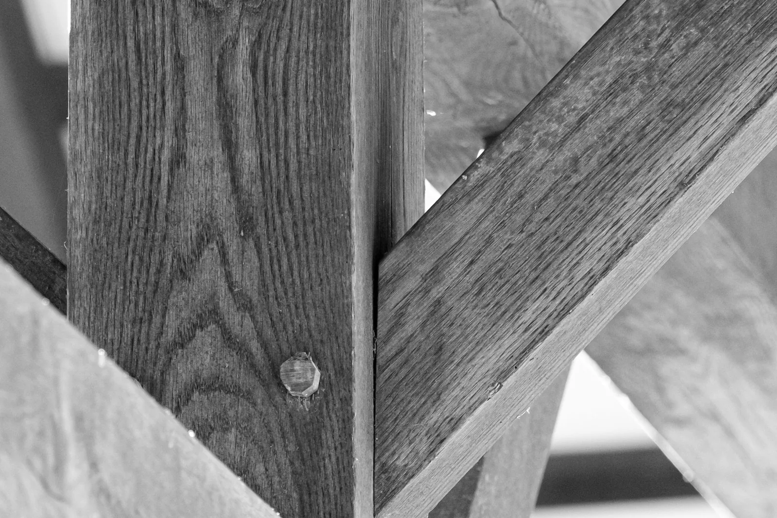 A weathered oak timber post with a round wooden peg driven flush through it, where two diagonal braces meet its faces along tightly cut joints.