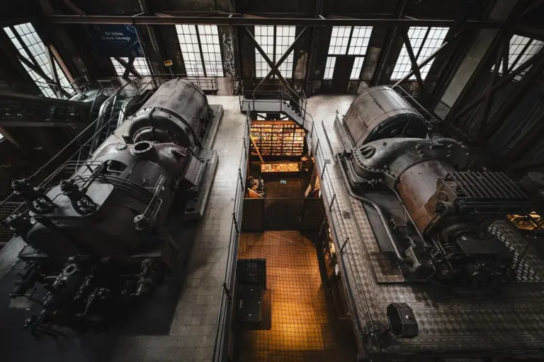 Two large historic engines standing side by side in a factory hall, seen from a walkway above, with daylight coming through a glazed roof.