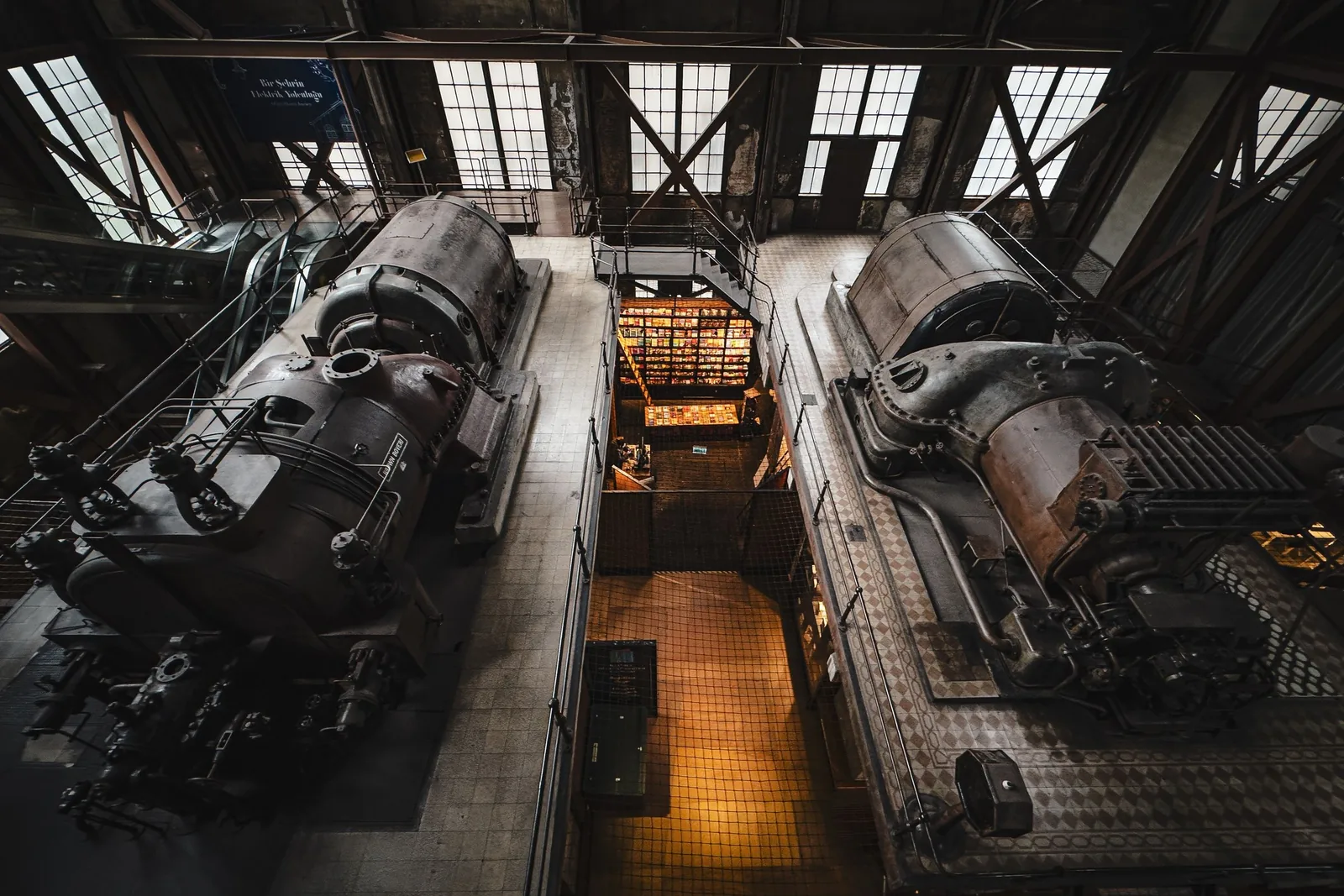 Two large historic engines standing side by side in a factory hall, seen from a walkway above, with daylight coming through a glazed roof.