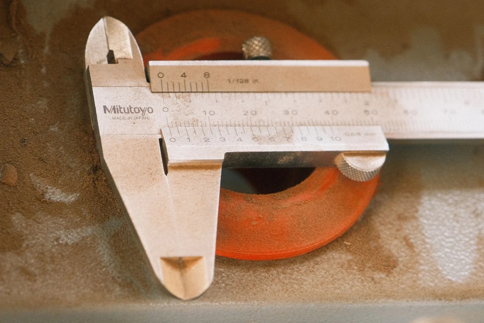 A metal vernier caliper resting on a worn workbench, its jaws set around a round orange object.