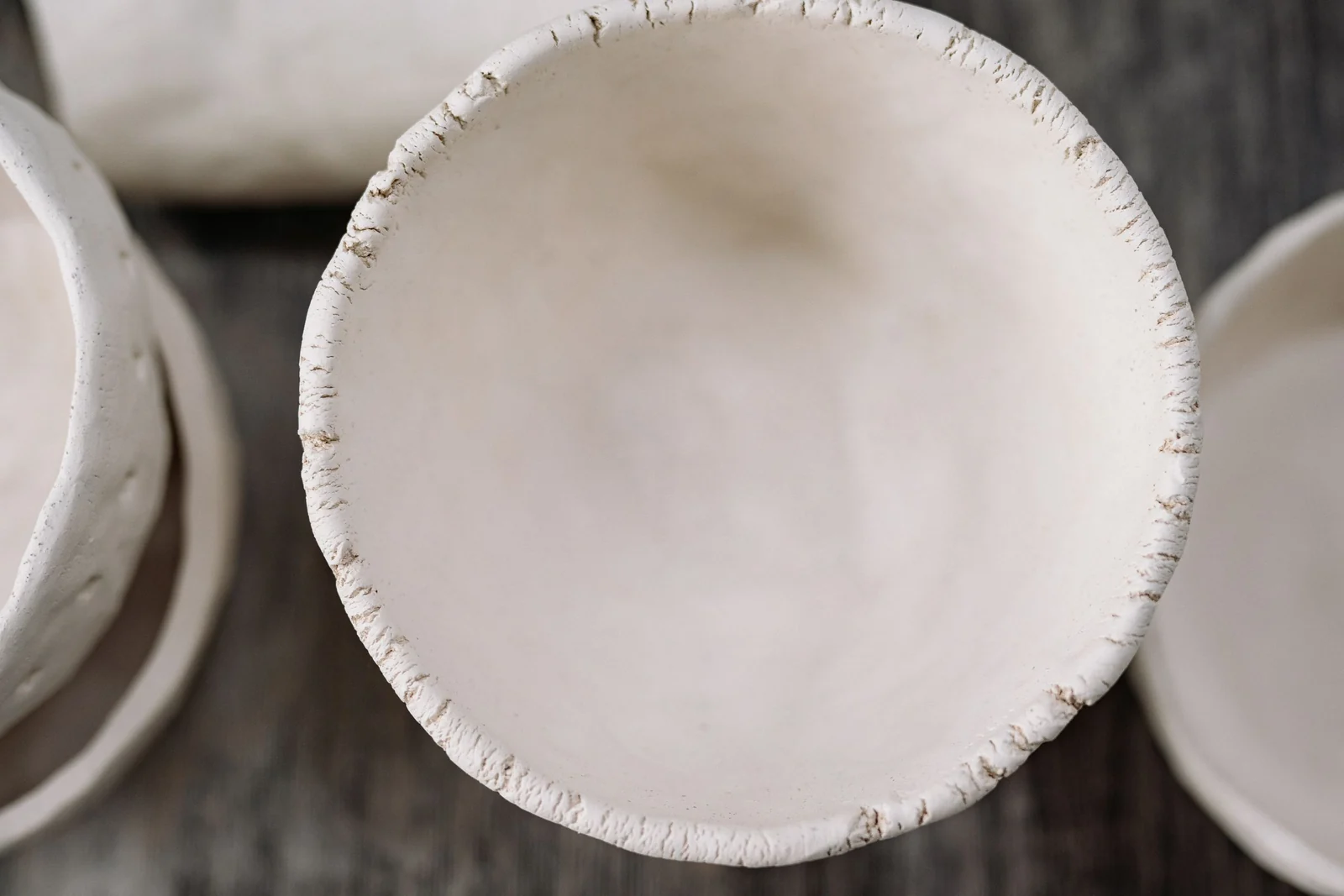 A handmade unglazed clay bowl seen from above, with an uneven hand-pinched rim, resting on a dark wooden surface.