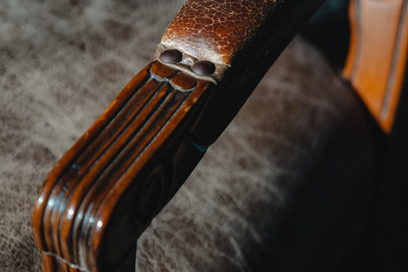 Close-up of an old wooden armchair, with a carved arm polished by handling and a cracked, worn leather armrest.