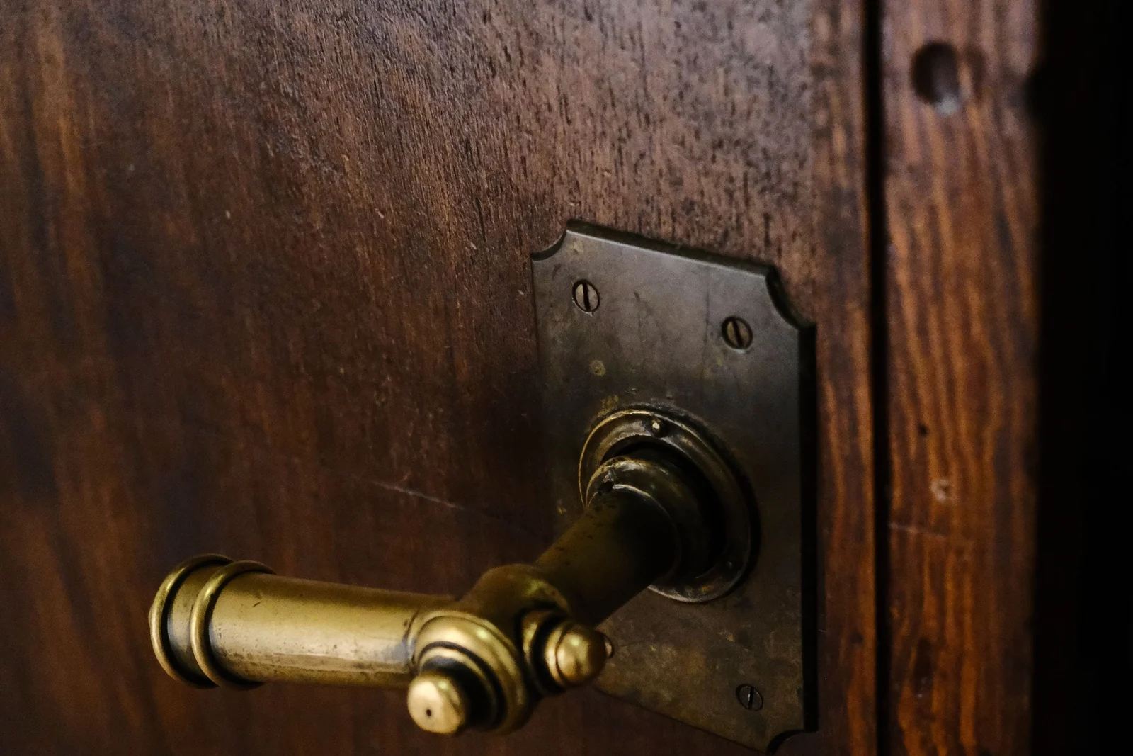 A brass door lever polished bright by constant use sits against the dark unpolished brass backplate on an aged wooden door.