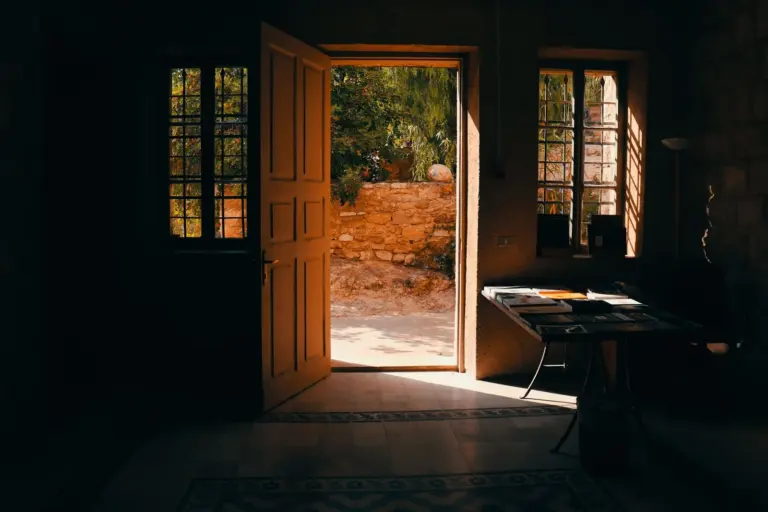 A pair of heavy wooden doors stands open in a dark room with bright sunlight falling on the courtyard beyond.