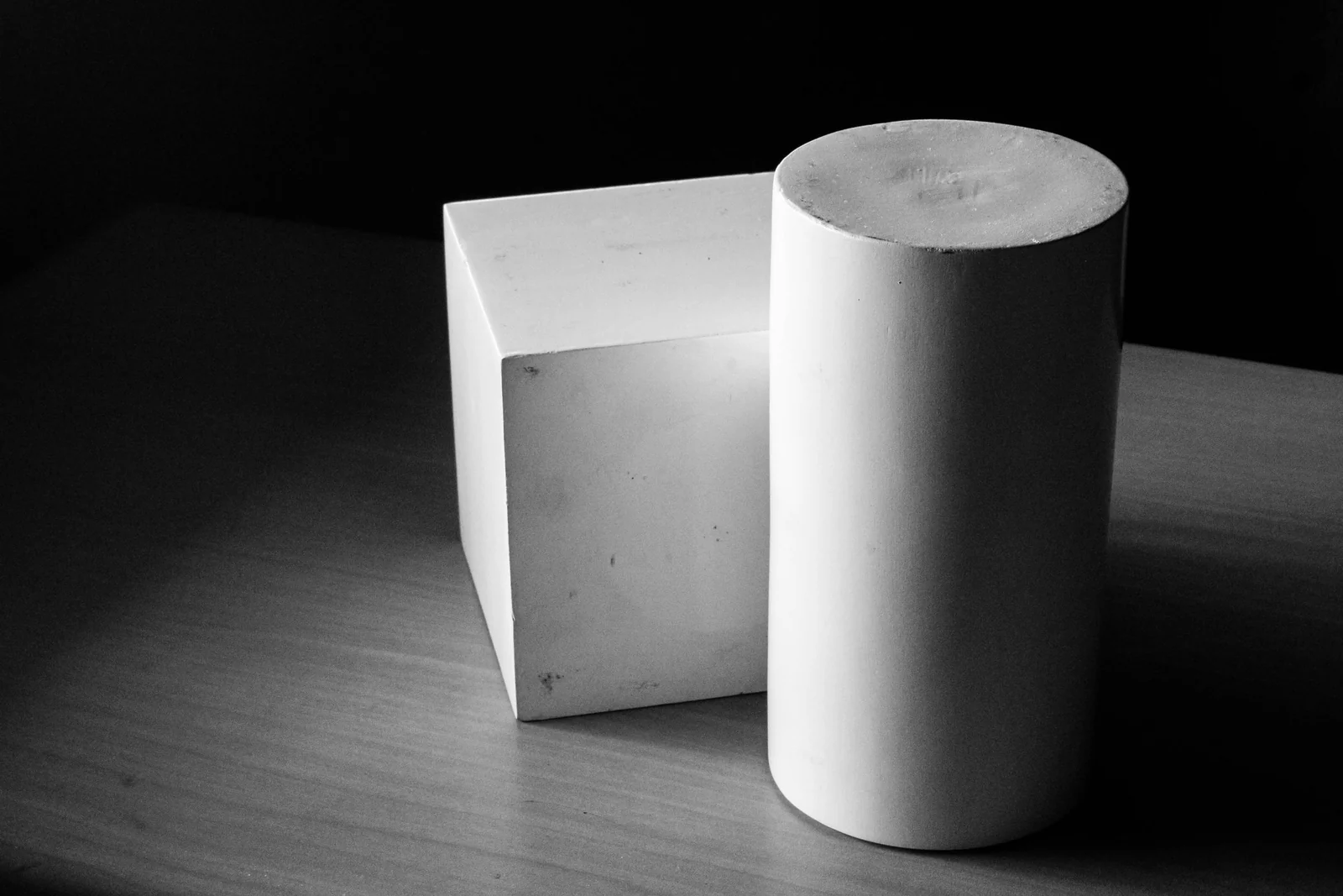 A plain cube and a plain cylinder standing apart from one another on a wooden table, lit from one side against a dark background, in black and white.