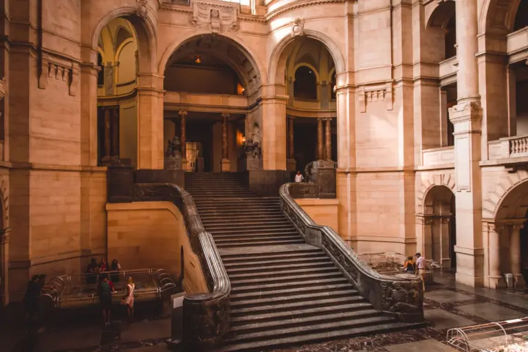 A wide sandstone hall with a broad staircase rising between arches; a few visitors stand at the far side, small in the frame.