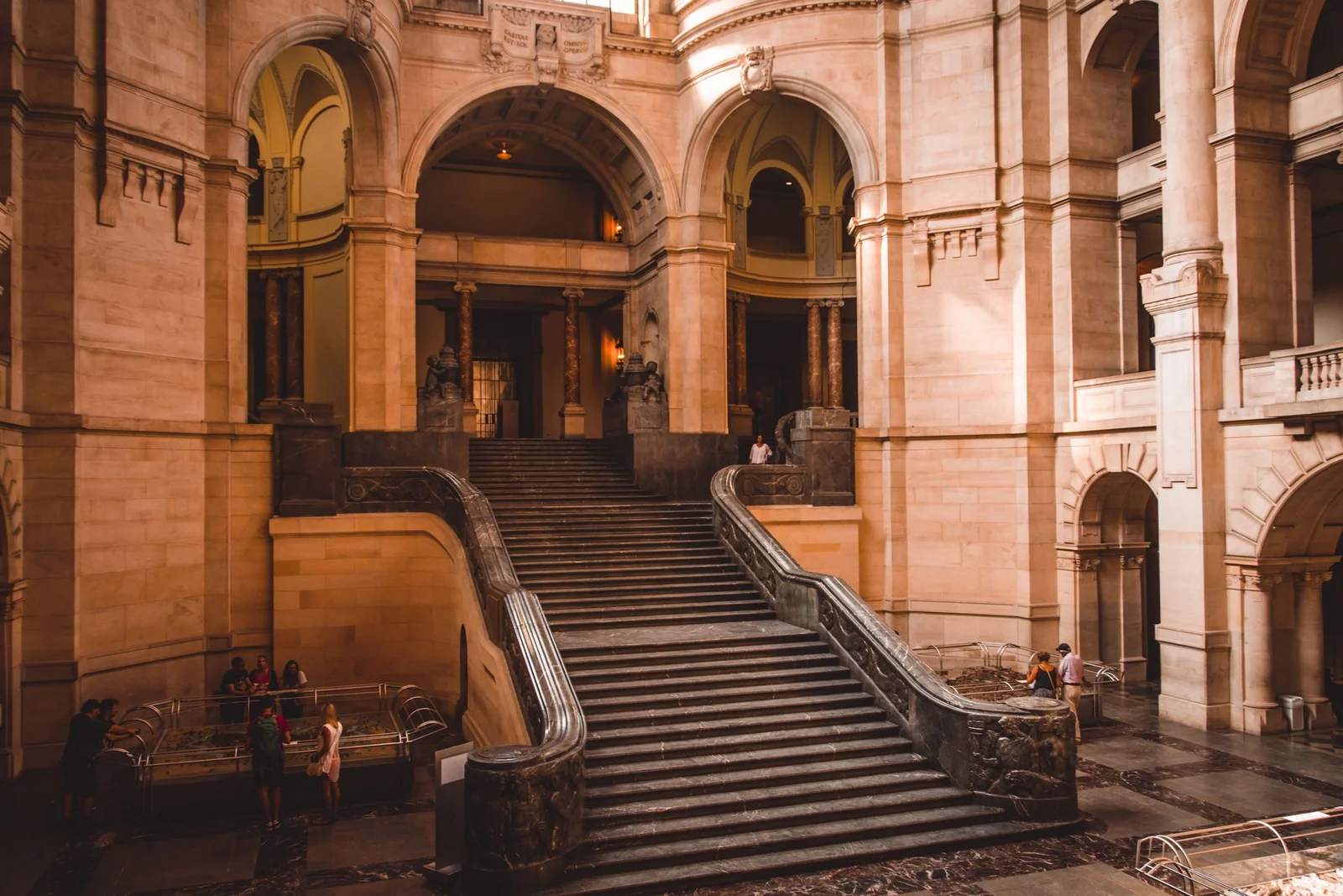 A wide sandstone hall with a broad staircase rising between arches; a few visitors stand at the far side, small in the frame.