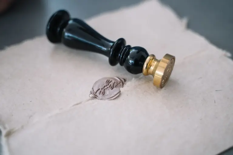 A brass and black-handled wax seal stamp lying on handmade paper beside a pressed wax impression.