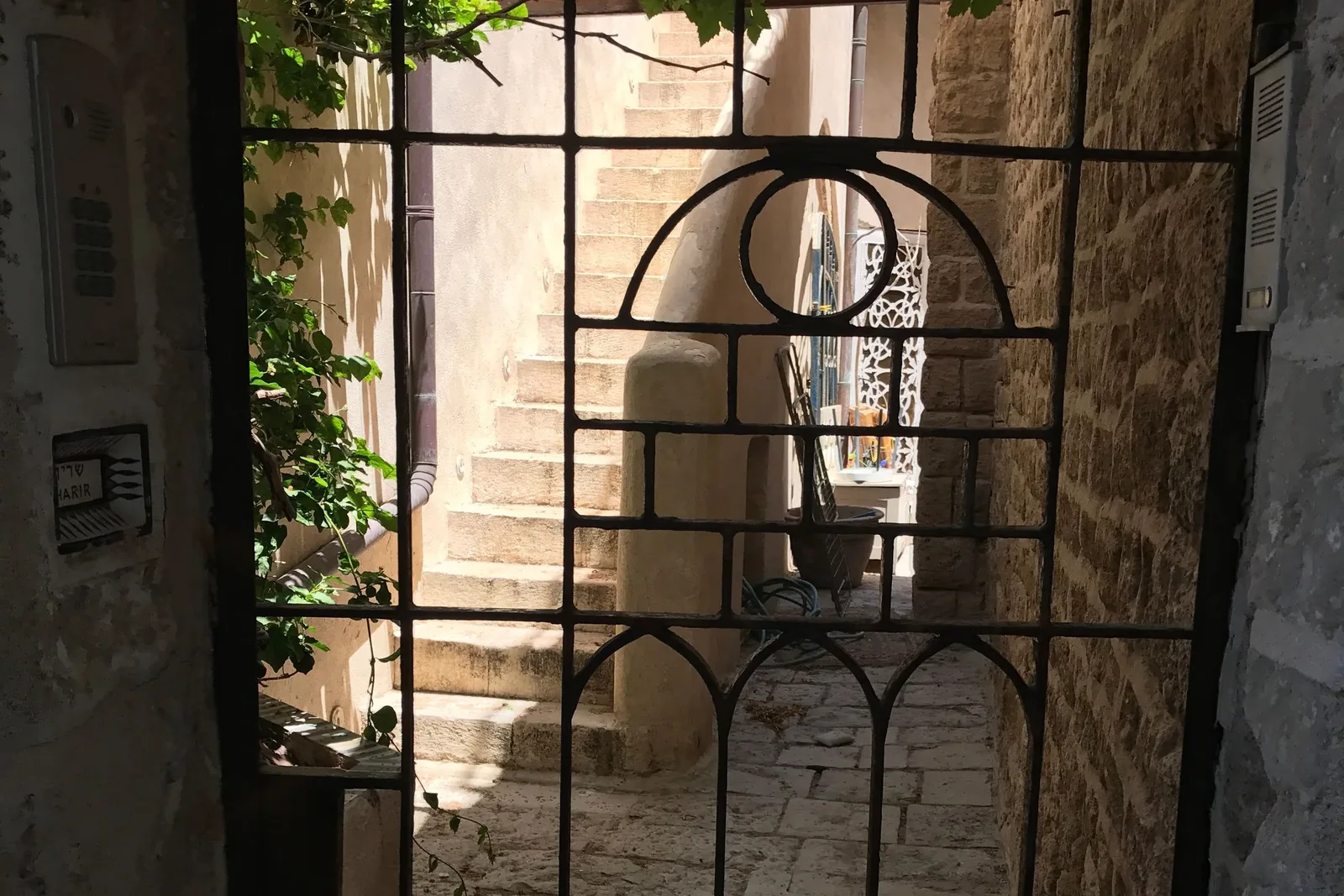 View through a wrought-iron gate into a narrow stone passage, with a staircase and daylight beyond.