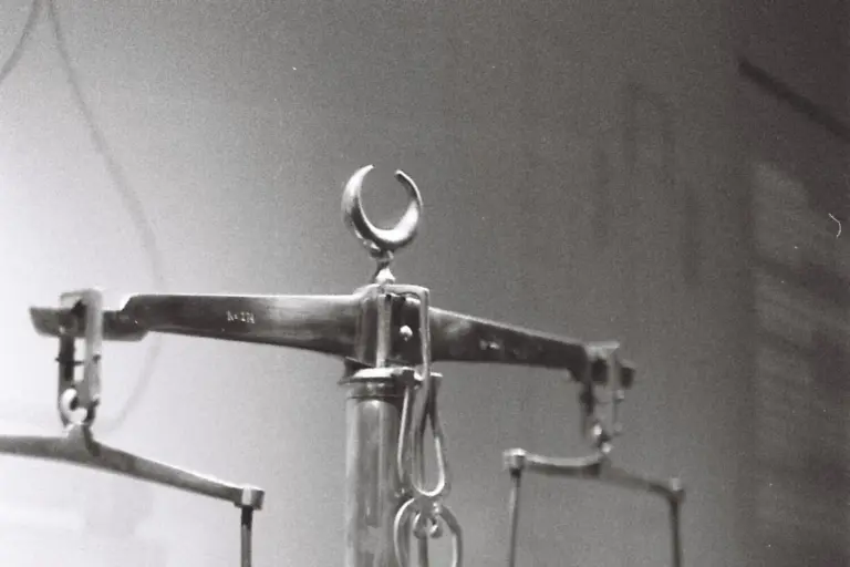 A vintage two-pan beam balance photographed in black and white, its pivot and suspension arms in sharp focus against a plain wall.