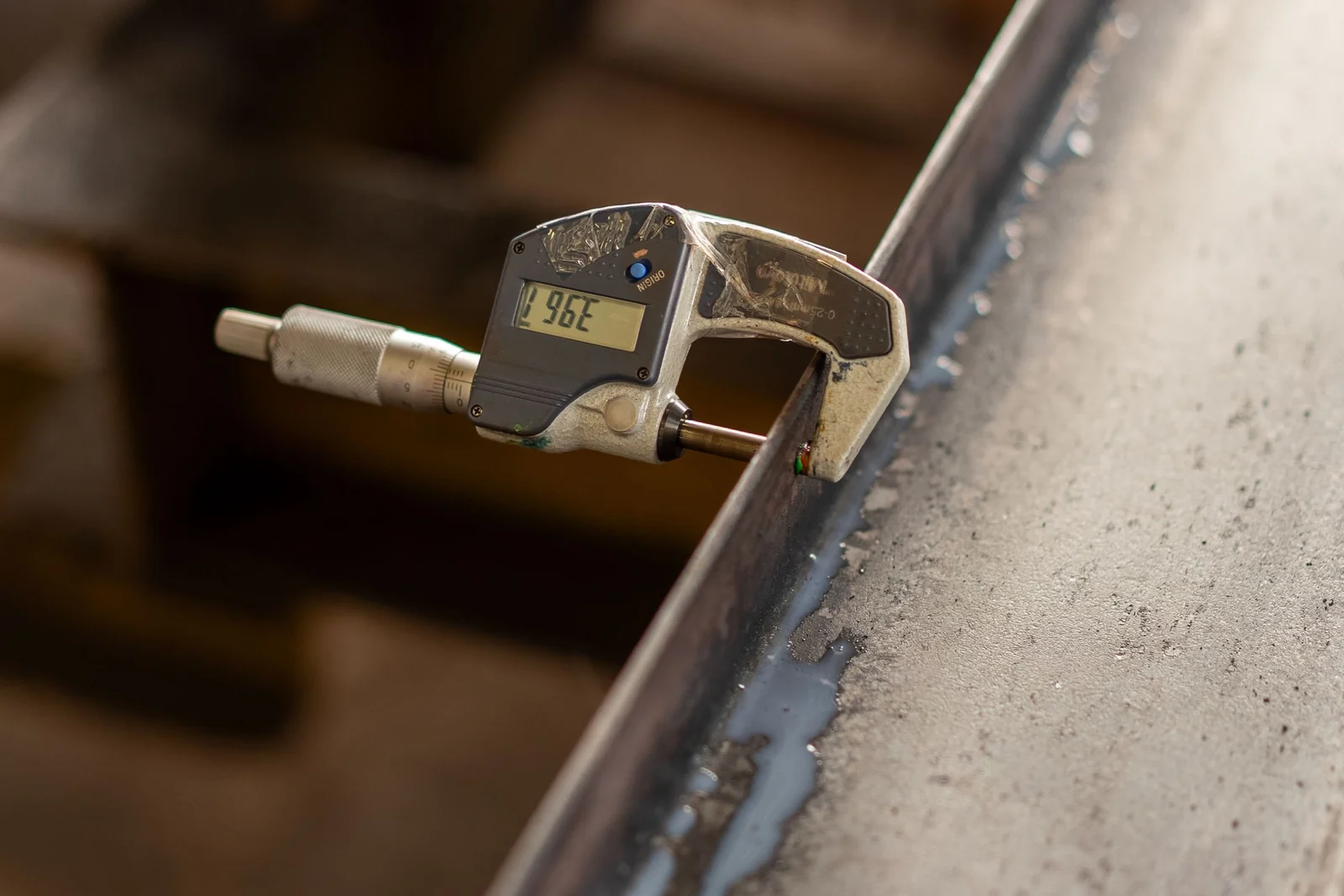 A micrometer clamped across the edge of a steel section on a workbench, its measuring faces closed on the metal.