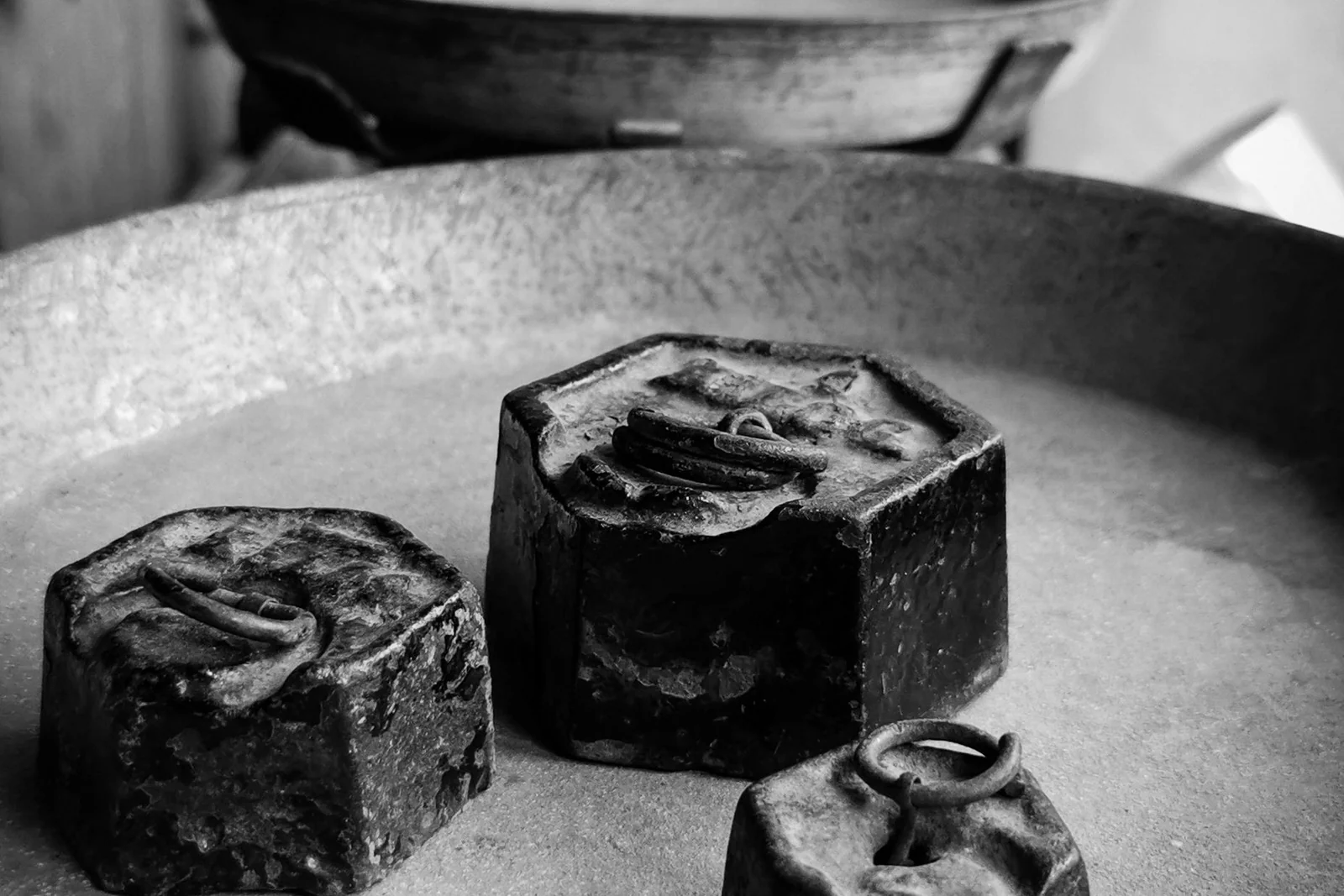 Three cast-iron weights of different sizes resting in the shallow pan of an old balance, photographed close up in black and white.