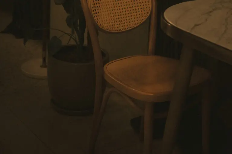 A single empty bentwood chair drawn up to a small marble-topped table in a warmly lit room, beside a potted plant.