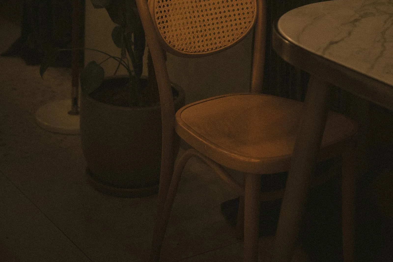 A single empty bentwood chair drawn up to a small marble-topped table in a warmly lit room, beside a potted plant.