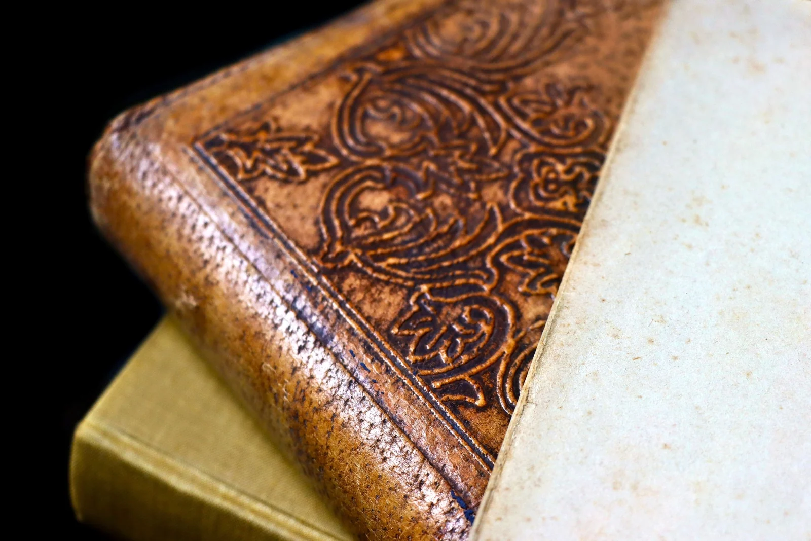 The corner of an antique leather-bound book with an ornate embossed border, resting on another volume.