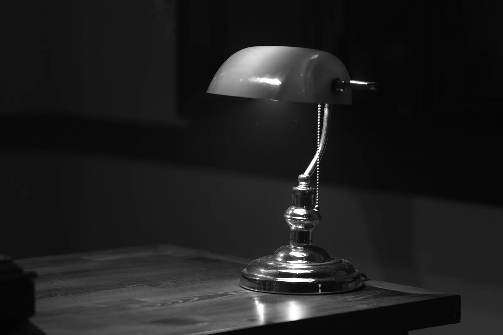 A banker's lamp lit on a wooden desk in an otherwise dark room, photographed in black and white, with papers beside it.