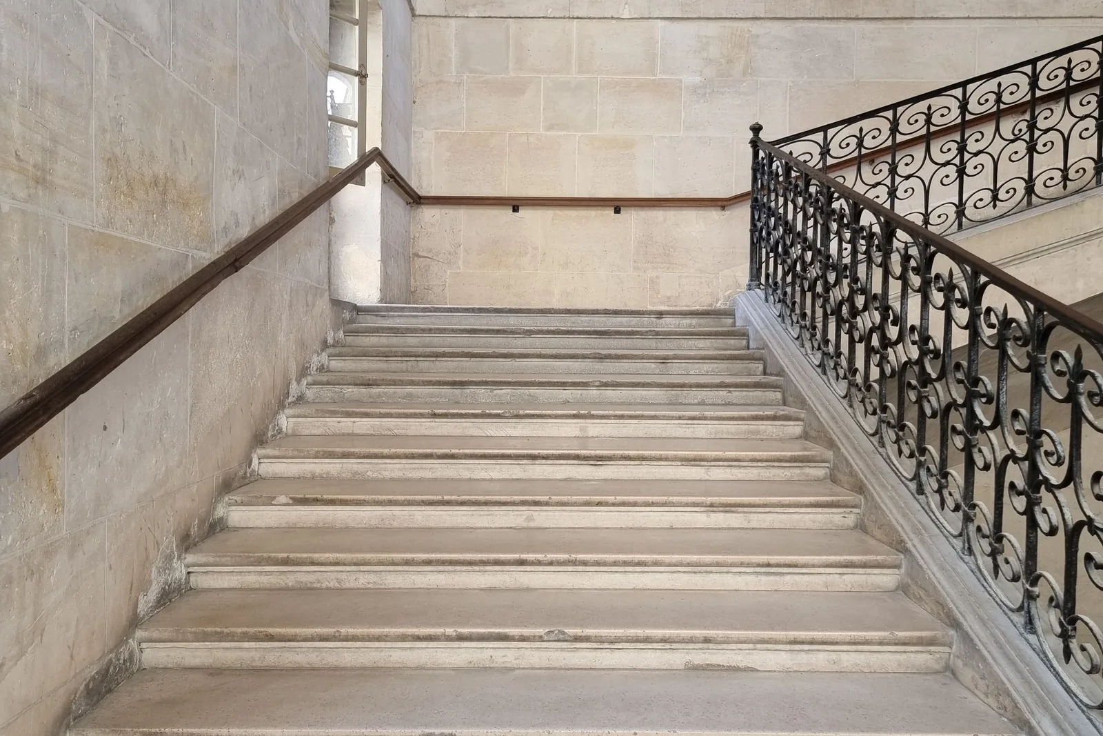 A straight flight of pale stone stairs rising between a plain wall and an ornate wrought-iron banister.