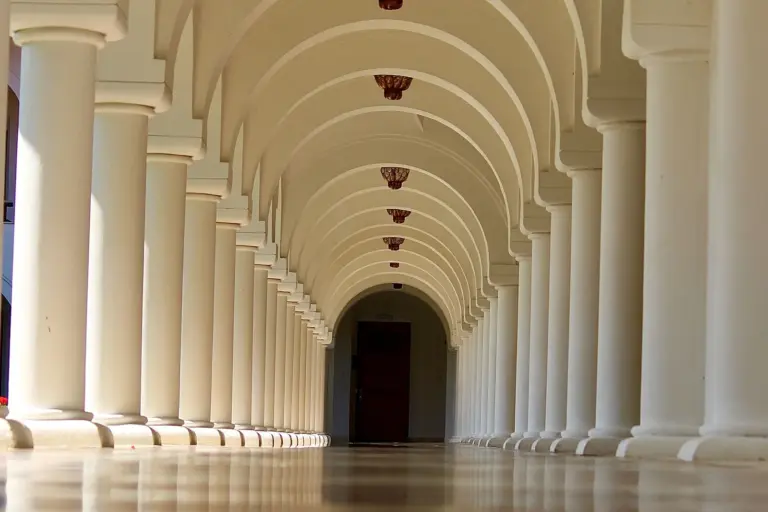 A long, empty vaulted corridor lined with tall white columns, receding towards a closed door at the far end.