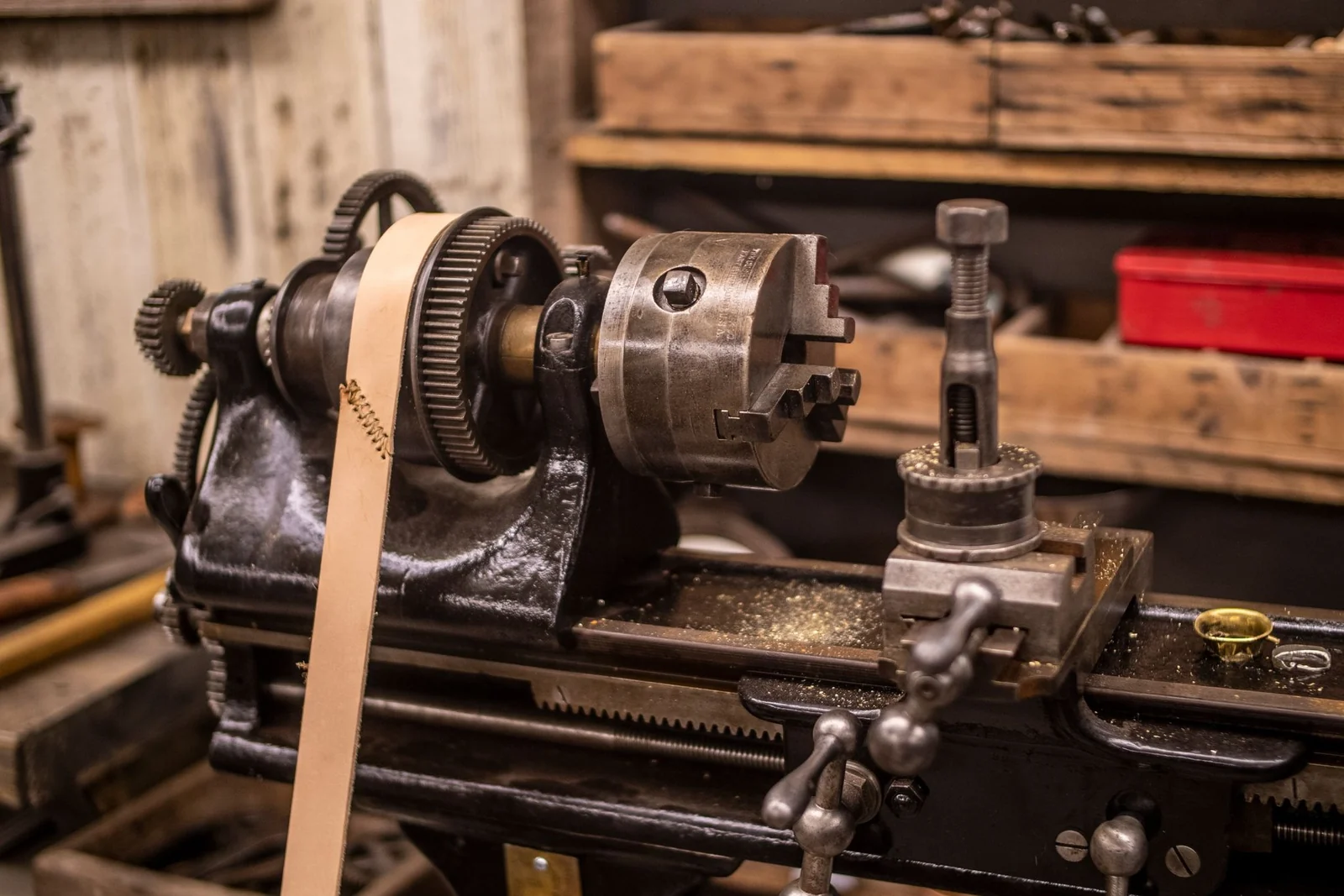 An antique lathe in a workshop, driven by a leather belt running over a pulley, with wooden shelves behind it.