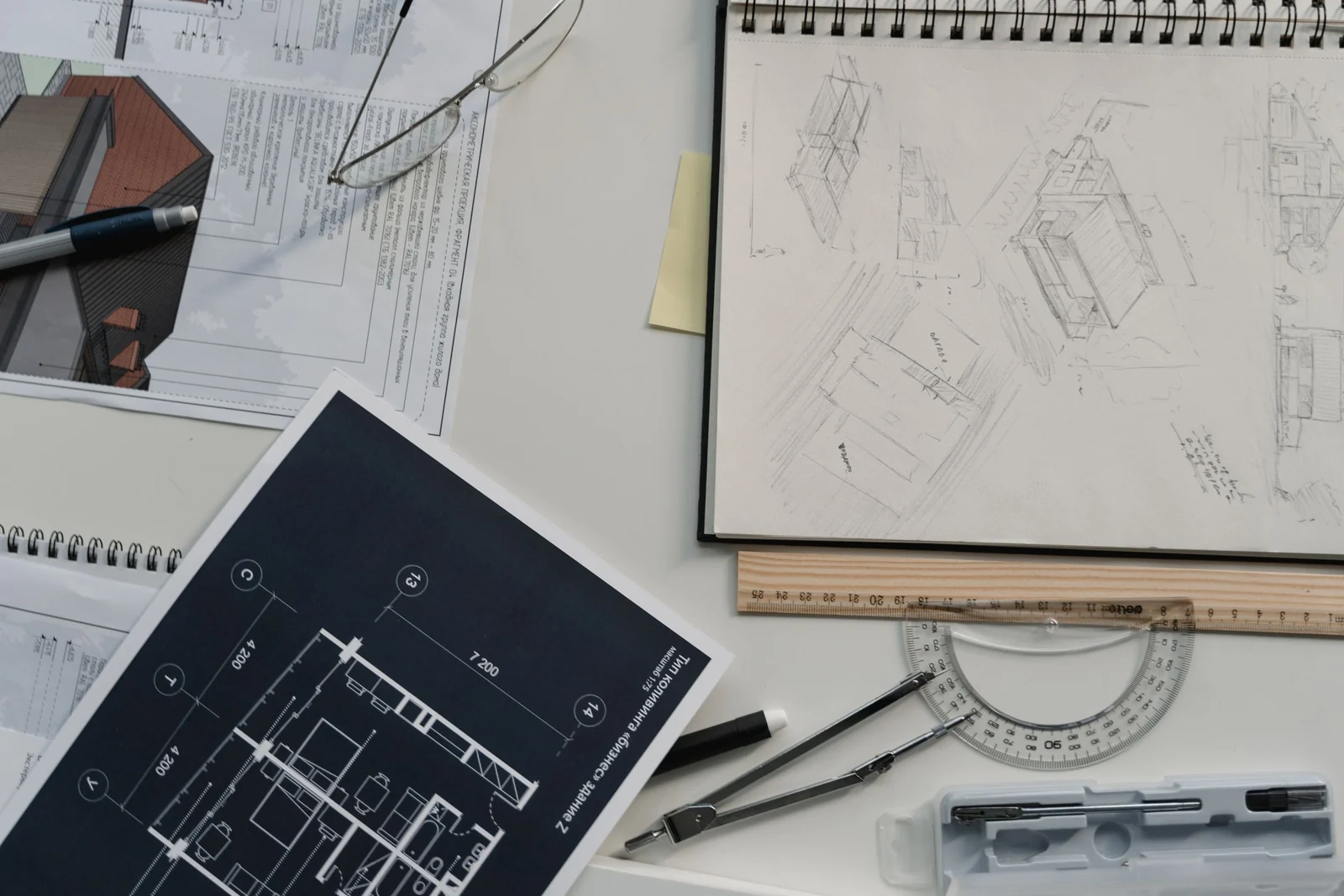A desk with a sketchbook of technical drawings, a printed plan, a set square, pencils and a pair of glasses.