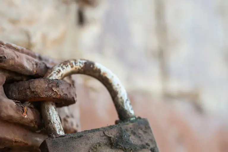 A heavy, rusted iron padlock hanging closed on a weathered metal hasp, photographed close up.