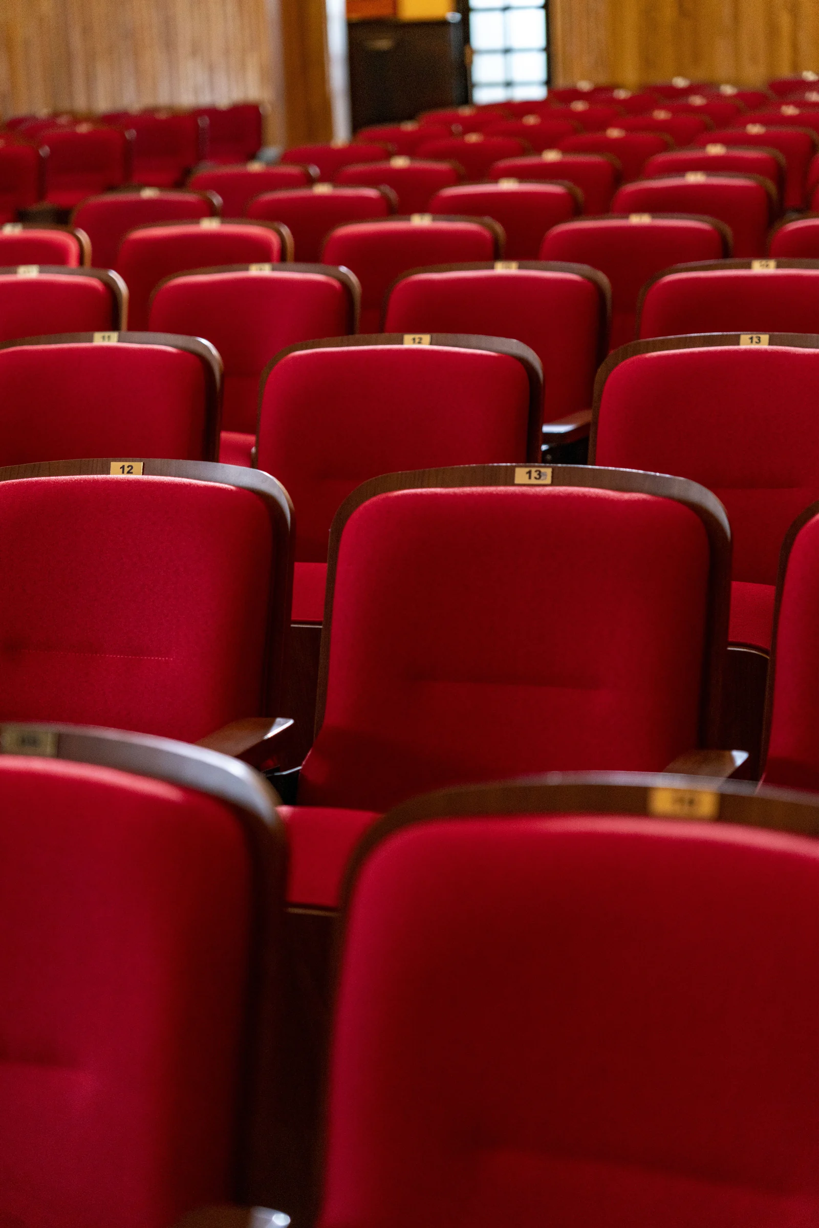 Rows of bright red theatre seats with wooden armrests and seat numbers, in an empty wood-panelled hall.