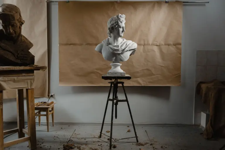 A white plaster bust stands on a sculptor's stand in a quiet studio, with a half-finished clay head on the workbench beside it.