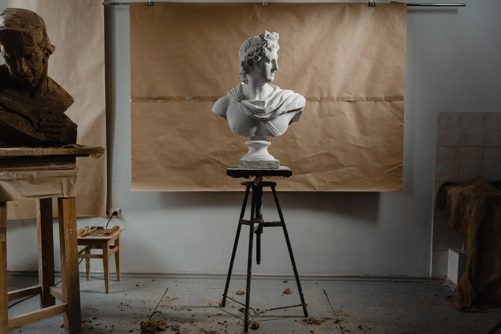 A white plaster bust stands on a sculptor's stand in a quiet studio, with a half-finished clay head on the workbench beside it.