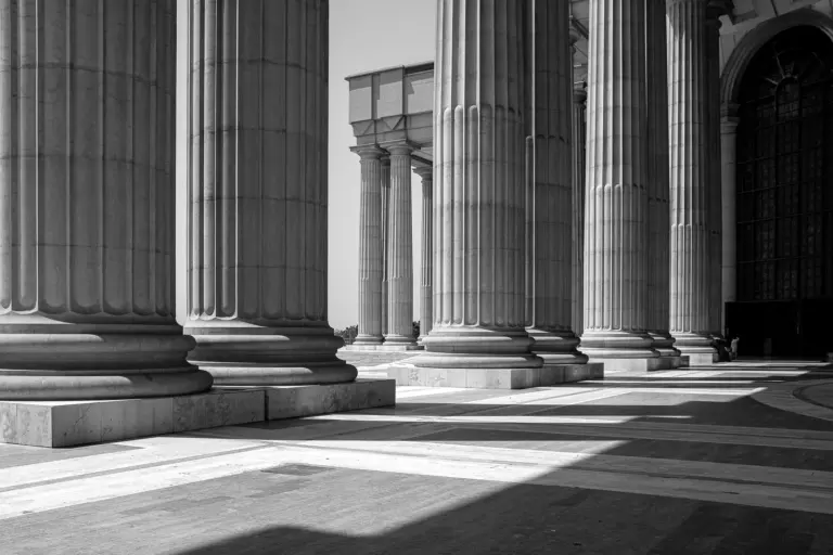 A black-and-white view of massive fluted stone columns on a paved forecourt, with sunlight falling between them.