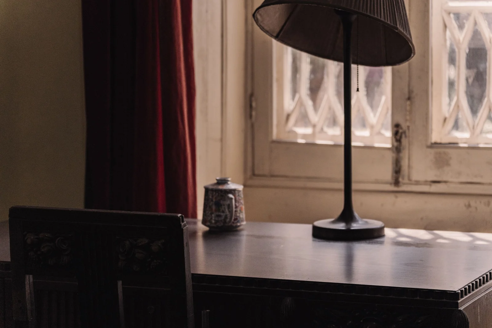A dark wooden desk with a tall lamp and a small porcelain jar, set before a window with carved lattice shutters.