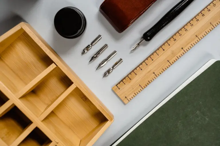 A wooden tray with empty compartments, a row of pen nibs, an ink pot and a ruler, laid out neatly on a desk.